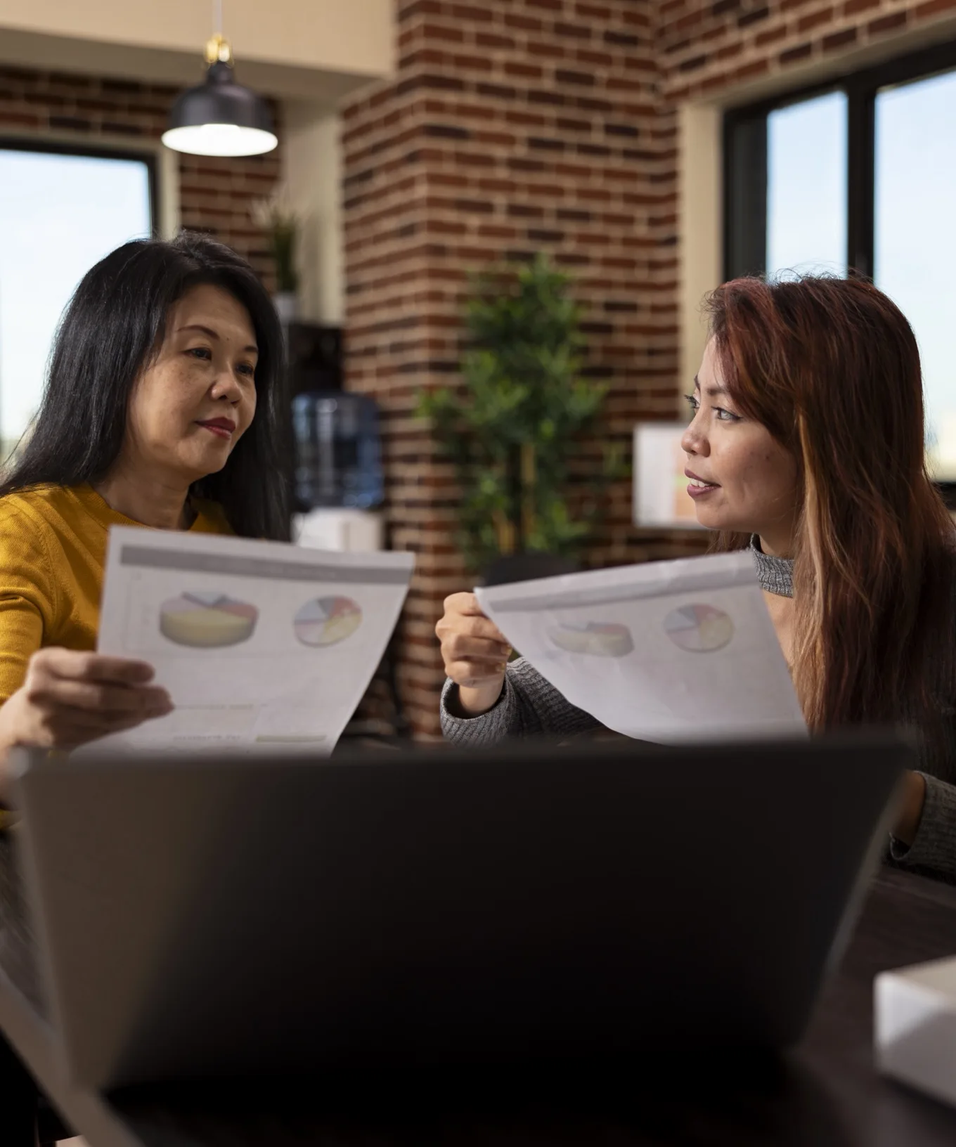Two colleagues in a brick-walled office holding printed charts and talking them through.